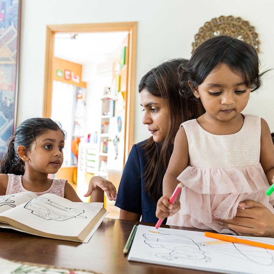 A woman and young girl, sitting at a table in a classroom, with a glue stick and paper while working together on an activity.