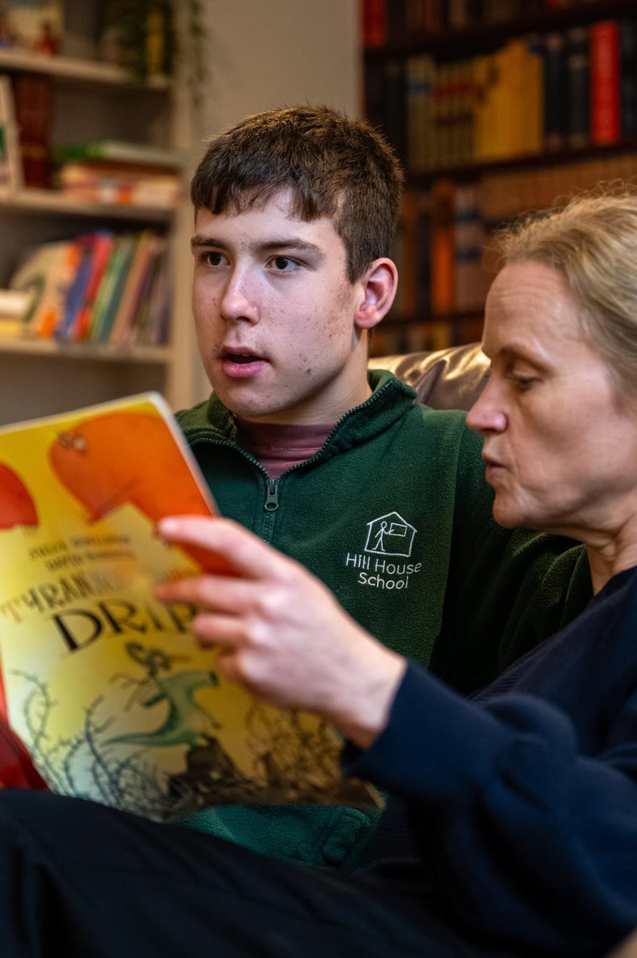 Young person and adult carer sitting together on a sofa, reading a large illustrated book in a cosy room with bookshelves in the background.