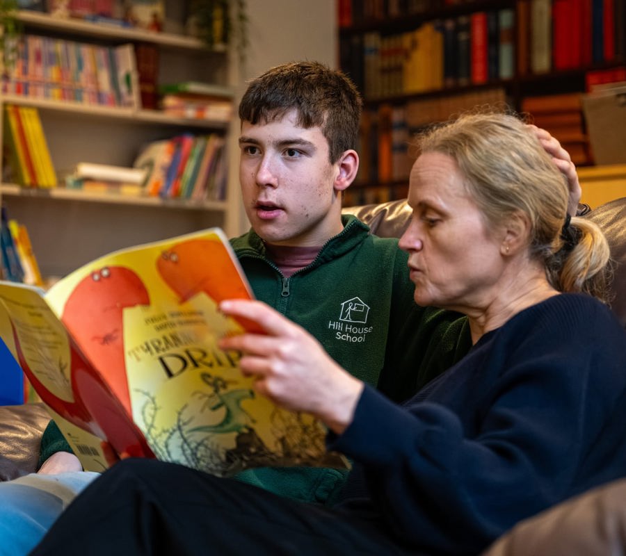 Young person and adult carer sitting together on a sofa, reading a large illustrated book in a cosy room with bookshelves in the background.