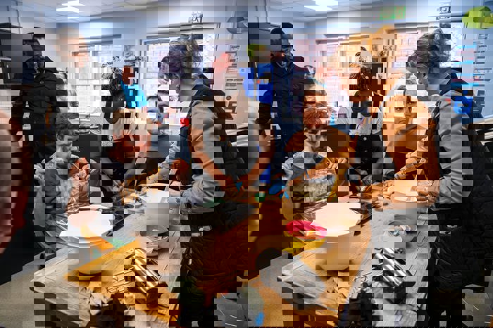 Group of adults gathered around a kitchen table preparing cupcakes, with staff supporting participants as they mix ingredients and fill baking trays.