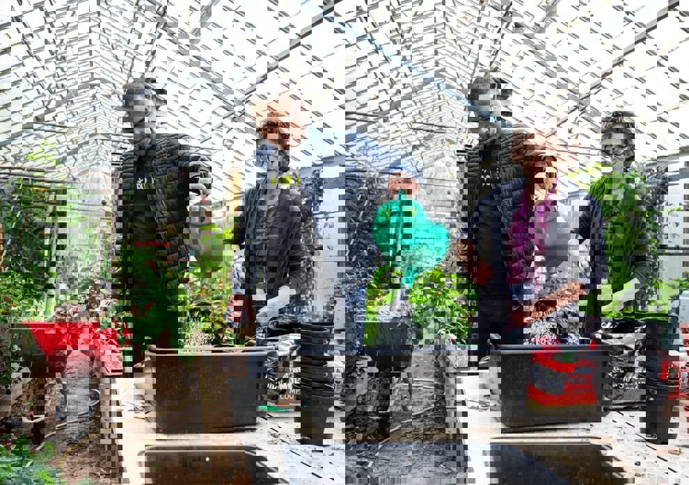 Young man watering plants in a greenhouse while a staff member supports him beside a potting table.