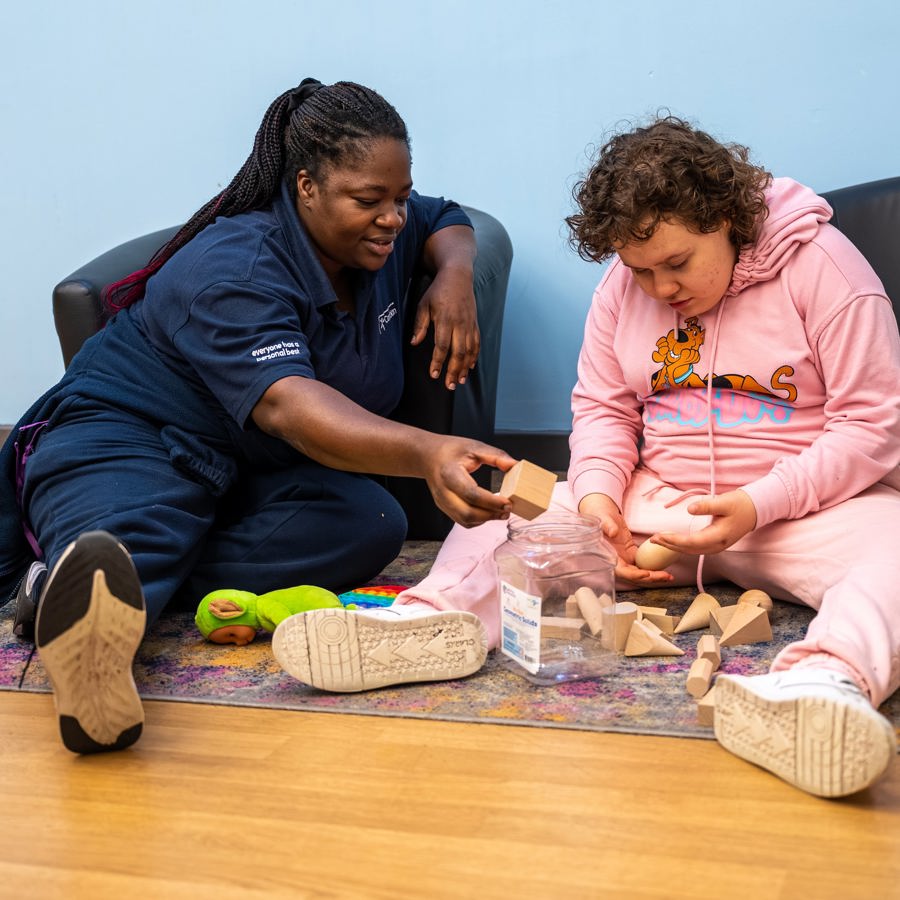 Carer sitting on the floor supporting a child as they explore and stack wooden blocks together.