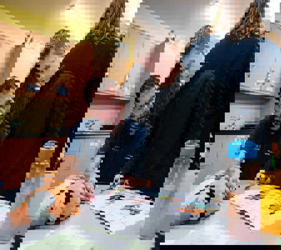 Adult supporting a child at a classroom table, pointing to a worksheet while the child follows along with a pencil.