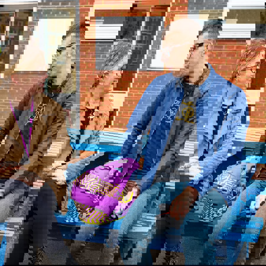 Two people sitting on a blue bench outside a brick building, chatting, with a purple helmet placed between them.