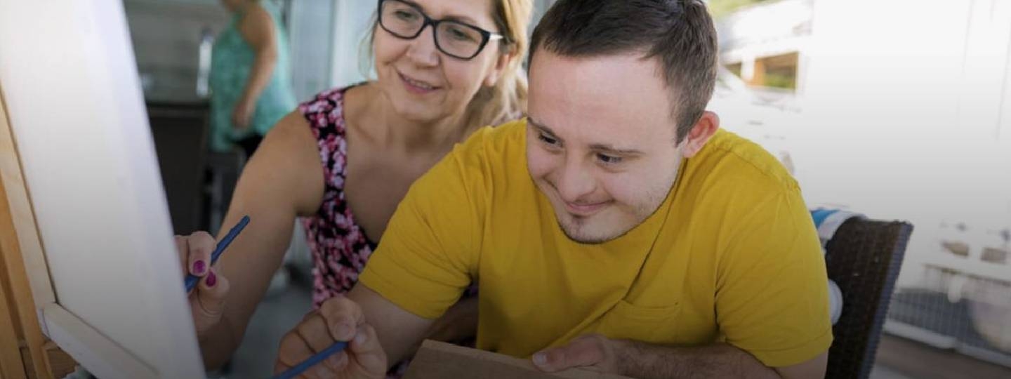 young man painting with his carer behind him.