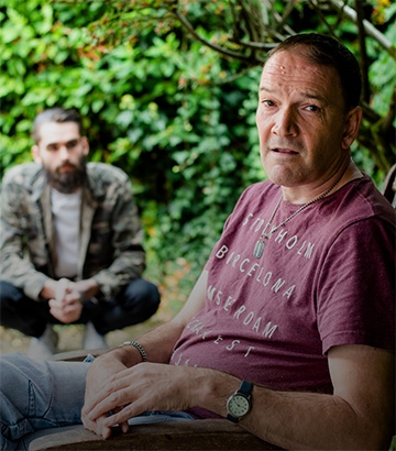 man sat in chair looking at the camera with his carer in the background.